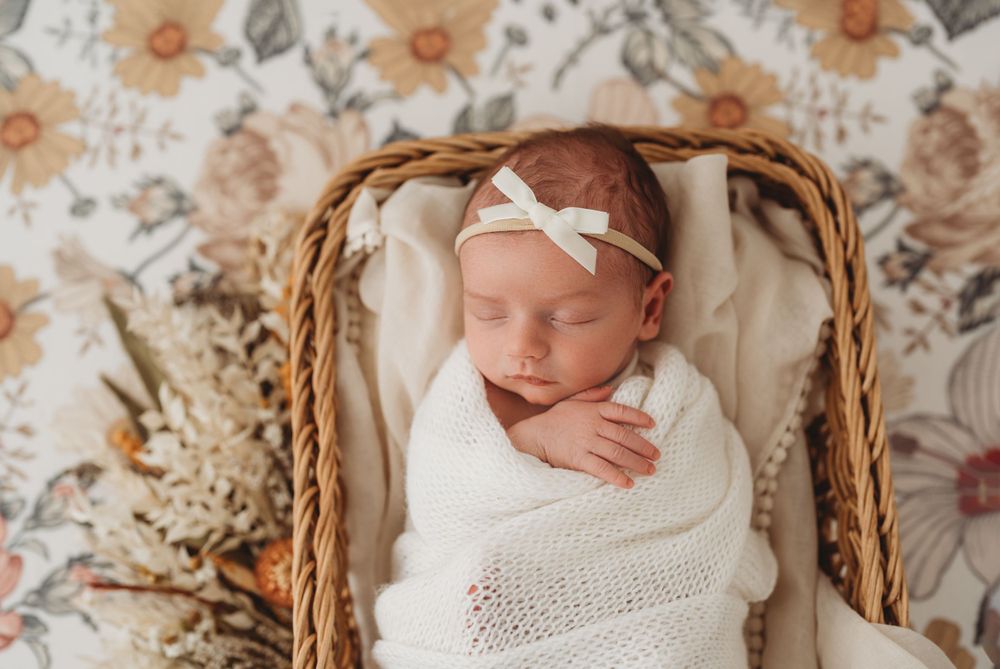 Sleeping newborn in white wrap posed in wicker basket on floral background.