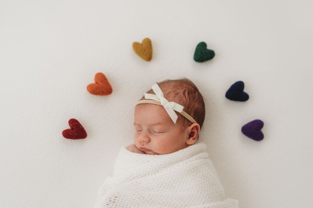 Sleeping newborn in white wrap surrounded by colorful heart shapes on white background.
