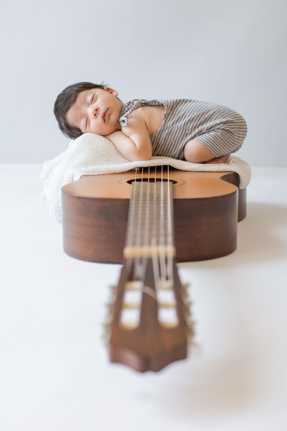 Sleeping newborn baby in striped outfit resting peacefully on acoustic guitar body with white background.