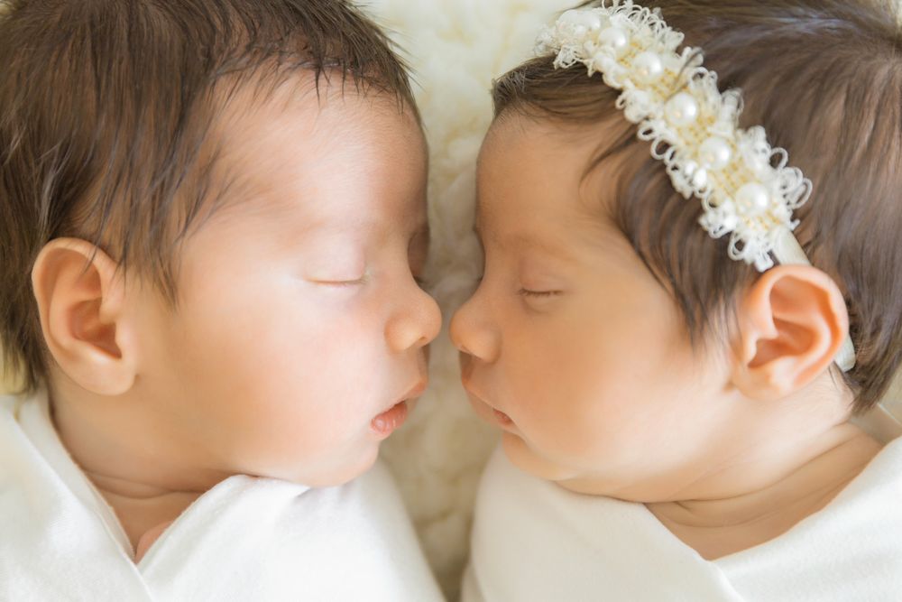Two sleeping newborn twins lying face to face, one wearing a pearl headband, dressed in white on a soft blanket.
