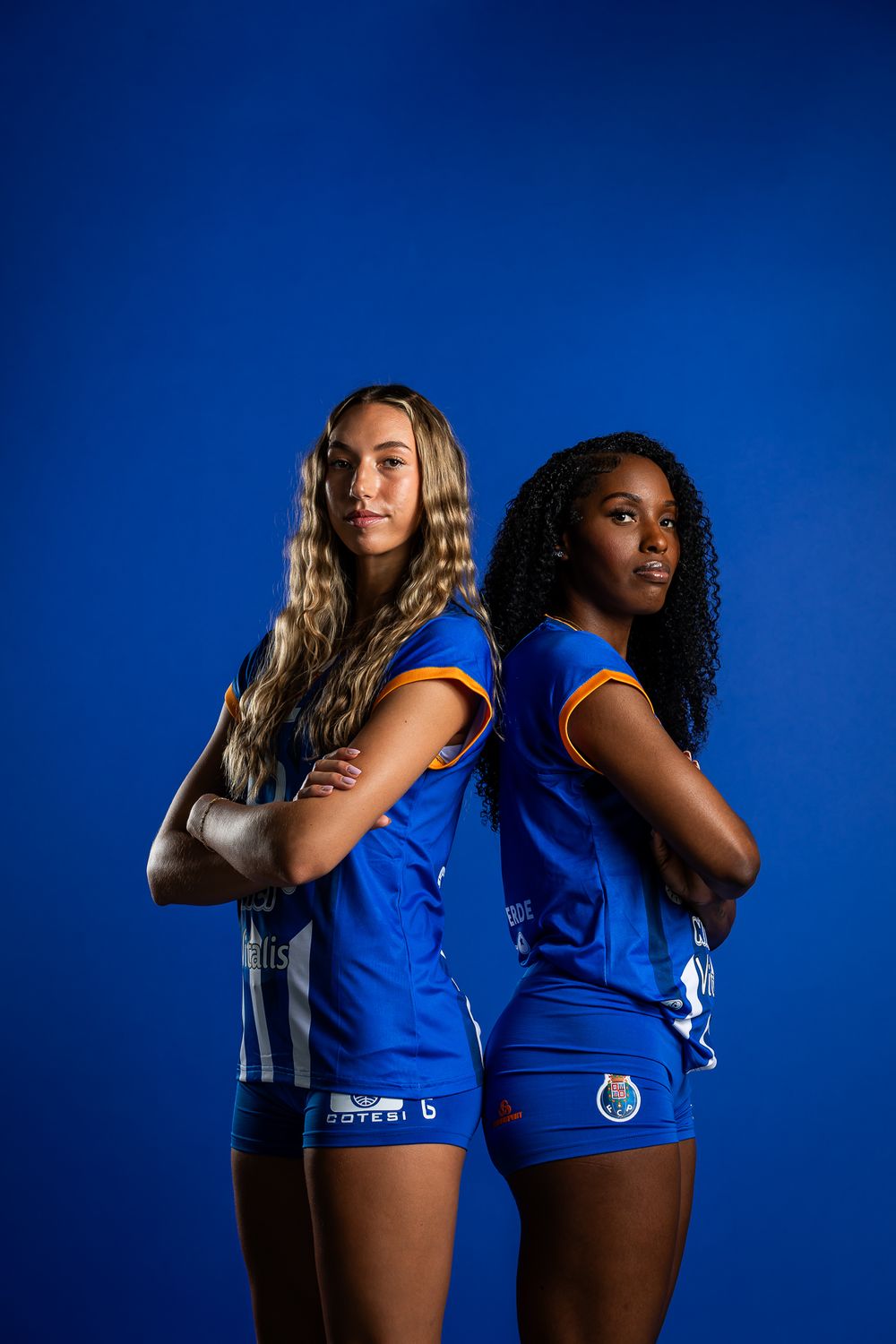 Two volleyball players in blue team uniforms pose back-to-back against a deep blue studio background.