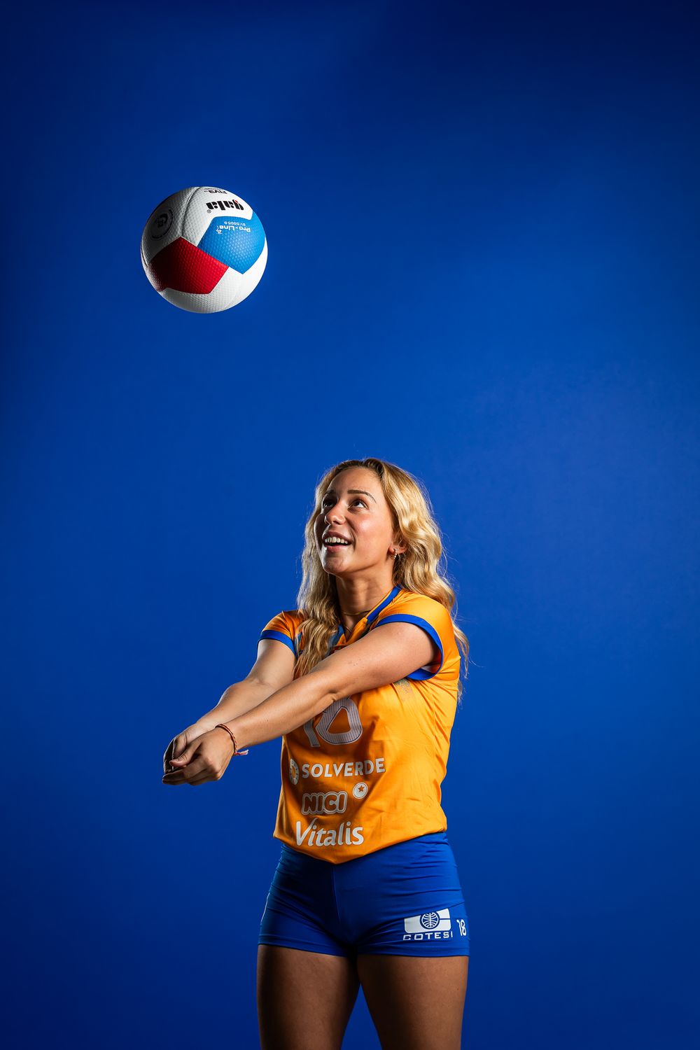 Volleyball player in yellow jersey and blue shorts bumps volleyball against vibrant blue background.