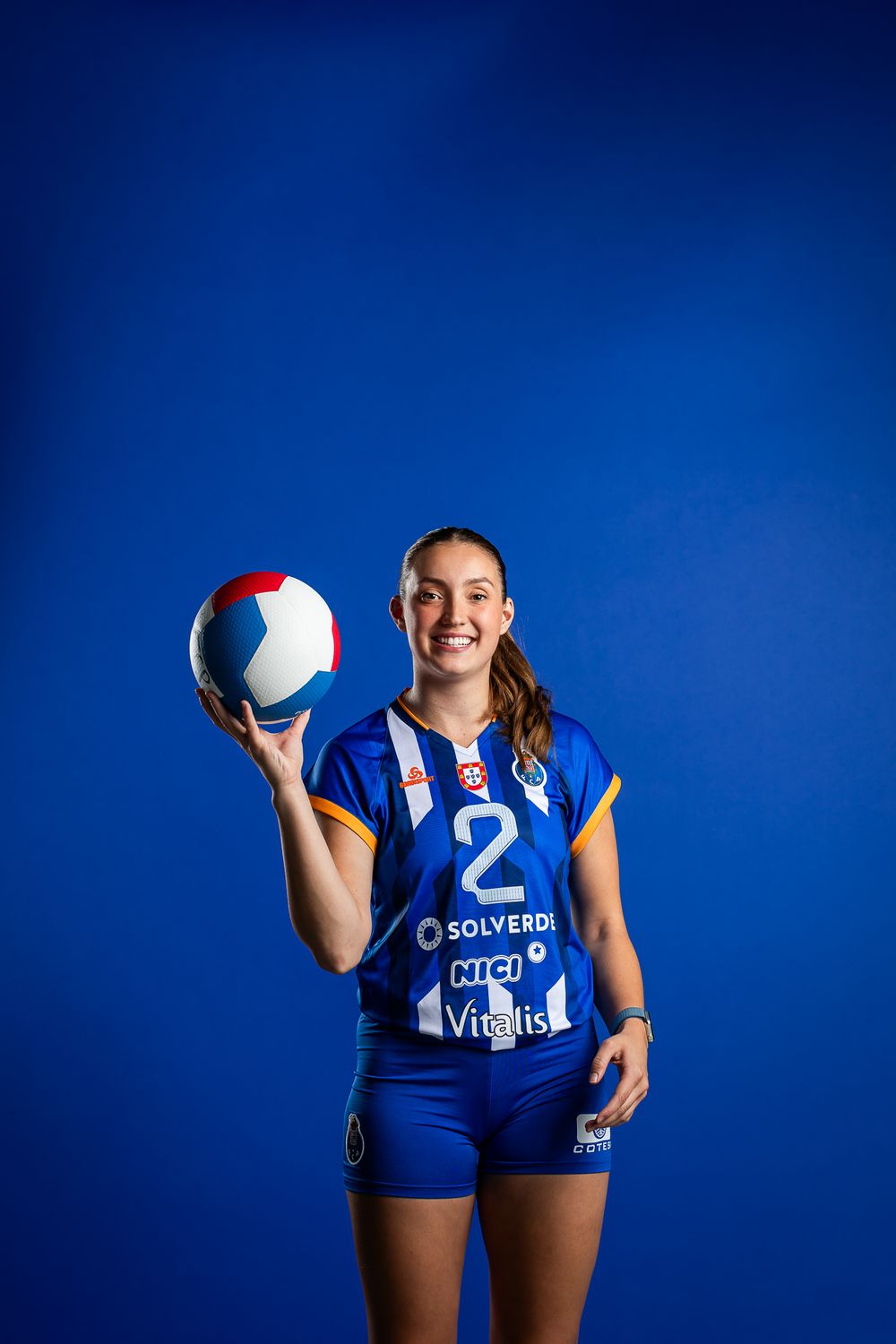 Volleyball athlete in blue uniform number 2 holds red and white ball while posing against bright blue background.