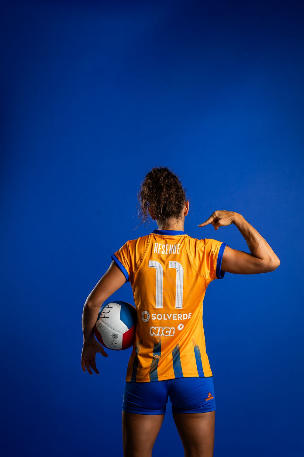 Sports player in yellow and blue striped uniform with number 11 poses with volleyball against blue background.