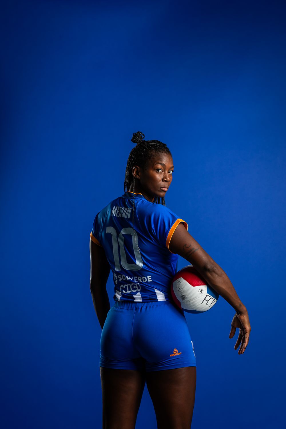 Professional athlete in blue volleyball uniform holding a ball poses against vibrant blue background for team photo shoot.