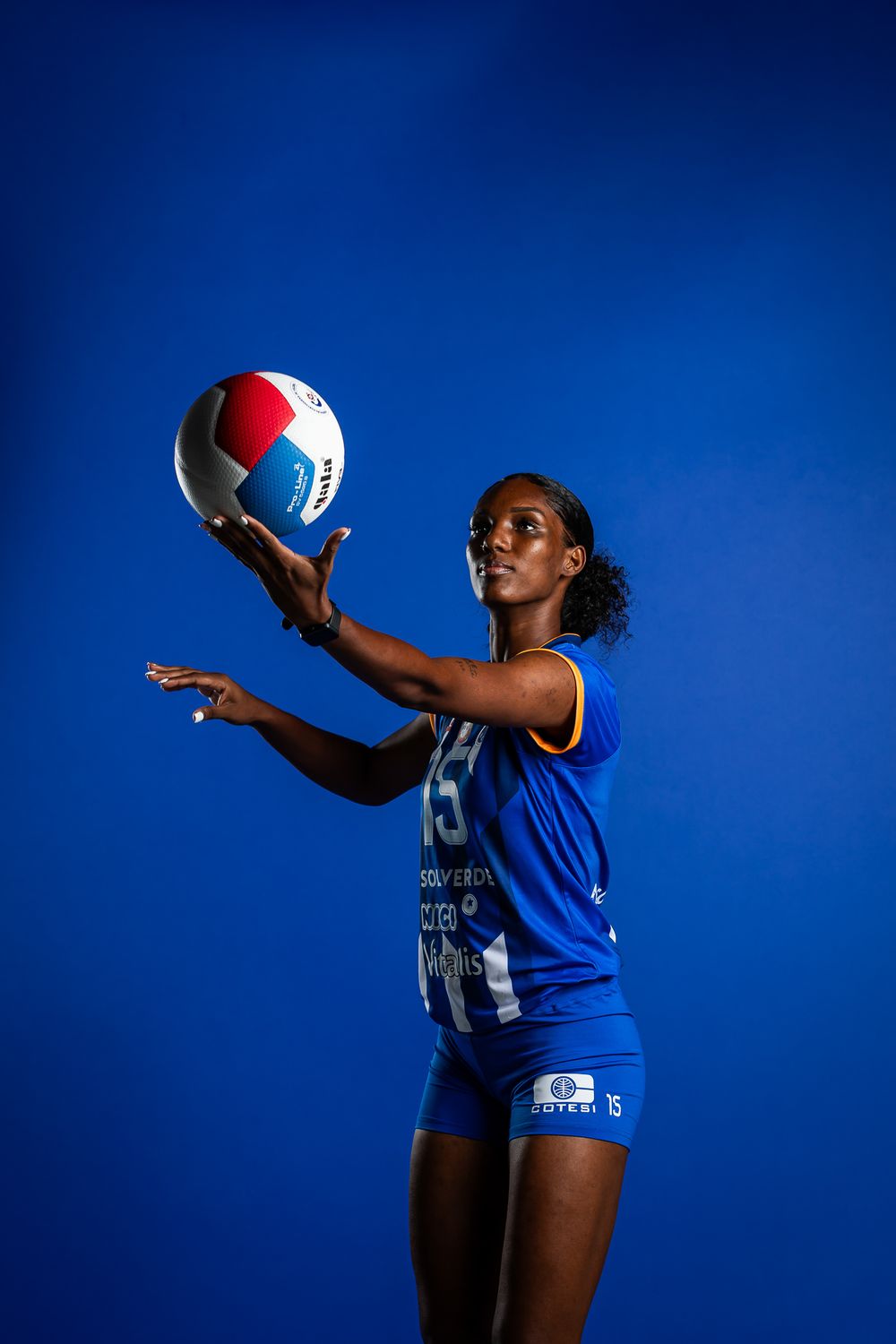 An athlete in a blue uniform holds up a volleyball against a deep blue background during a sports photo shoot.