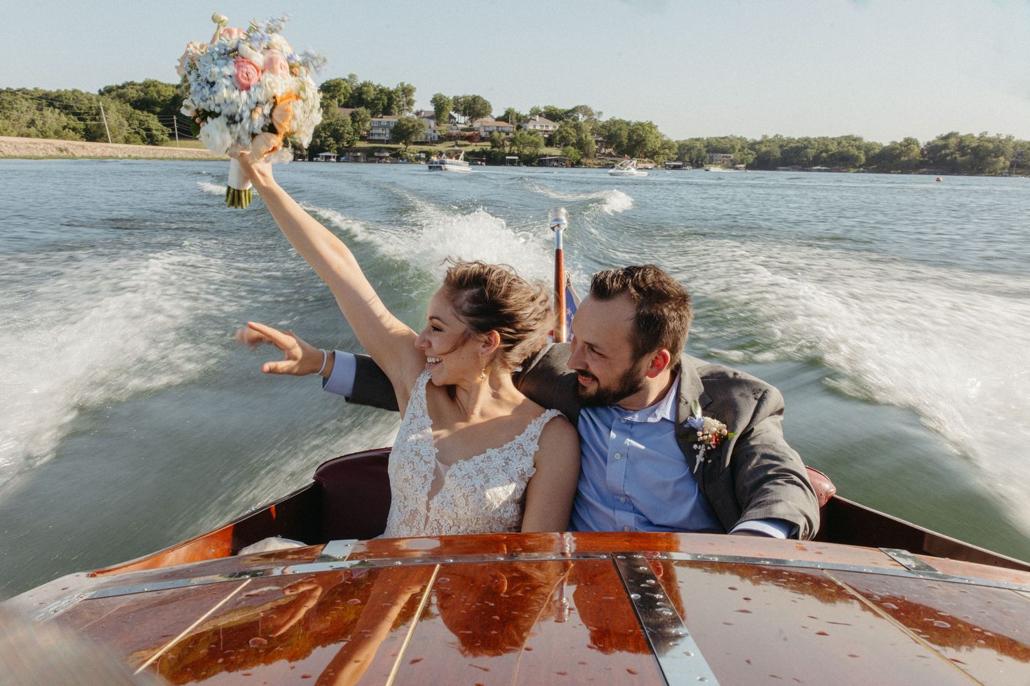 A couple celebrates on a wooden speedboat, holding a bouquet high while cruising across a lake.