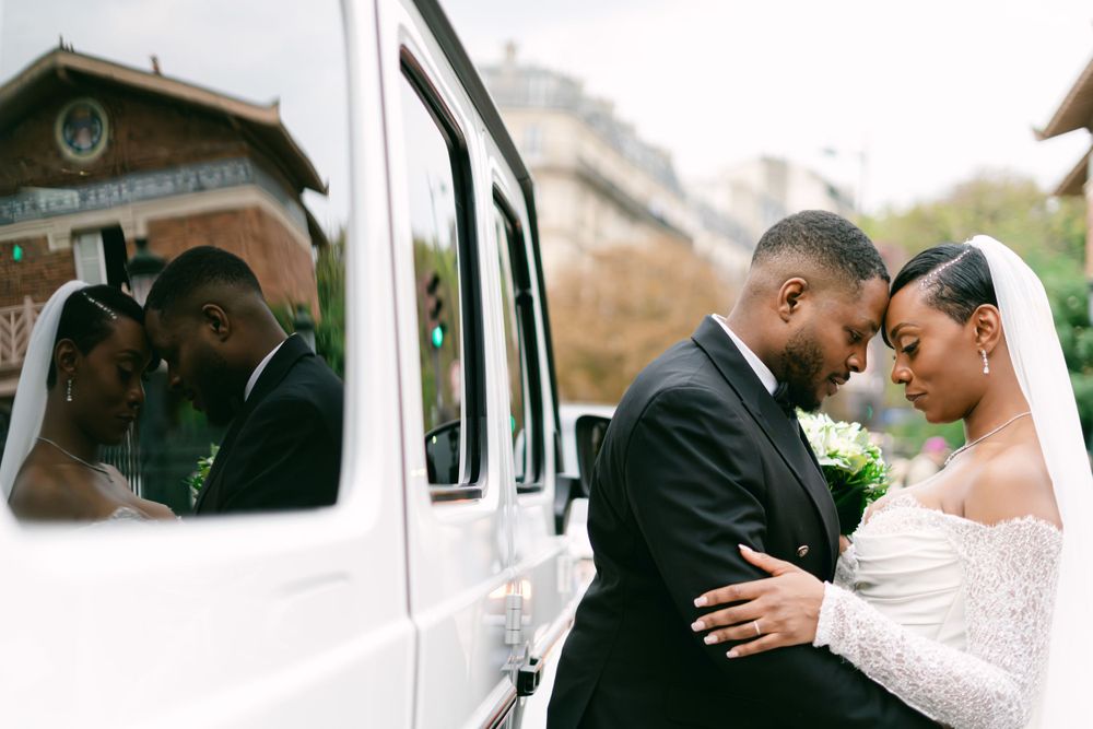 A couple shares an intimate moment beside a white vehicle on their wedding day while their reflection shows in the car window.