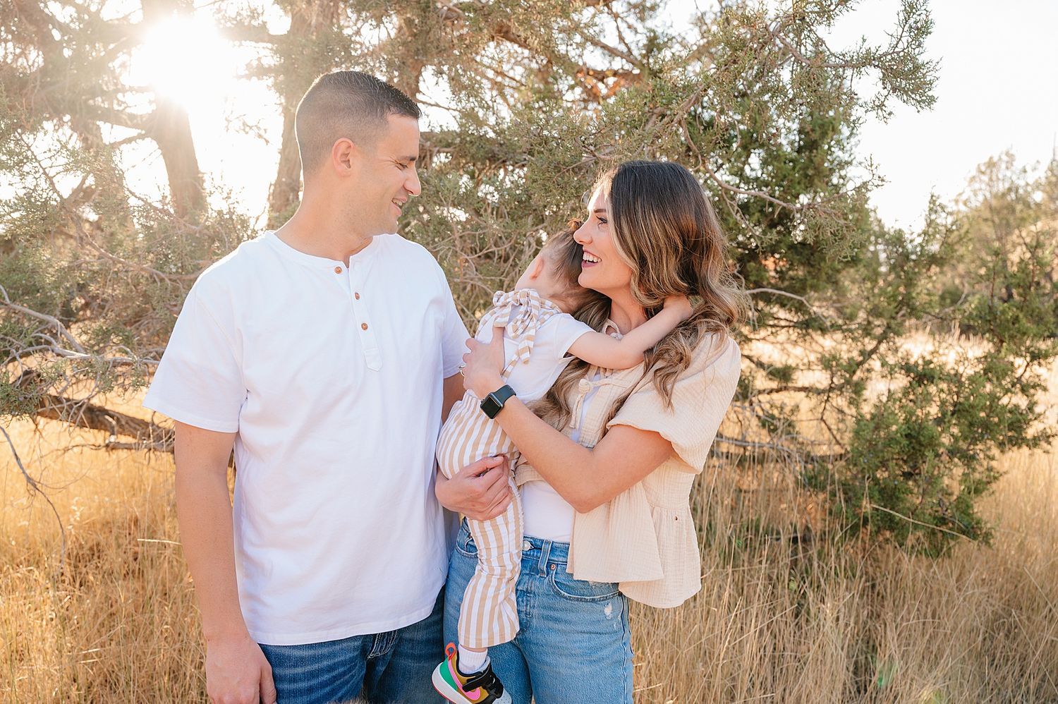 A couple and baby pose together in golden grass during sunset for a family portrait session.