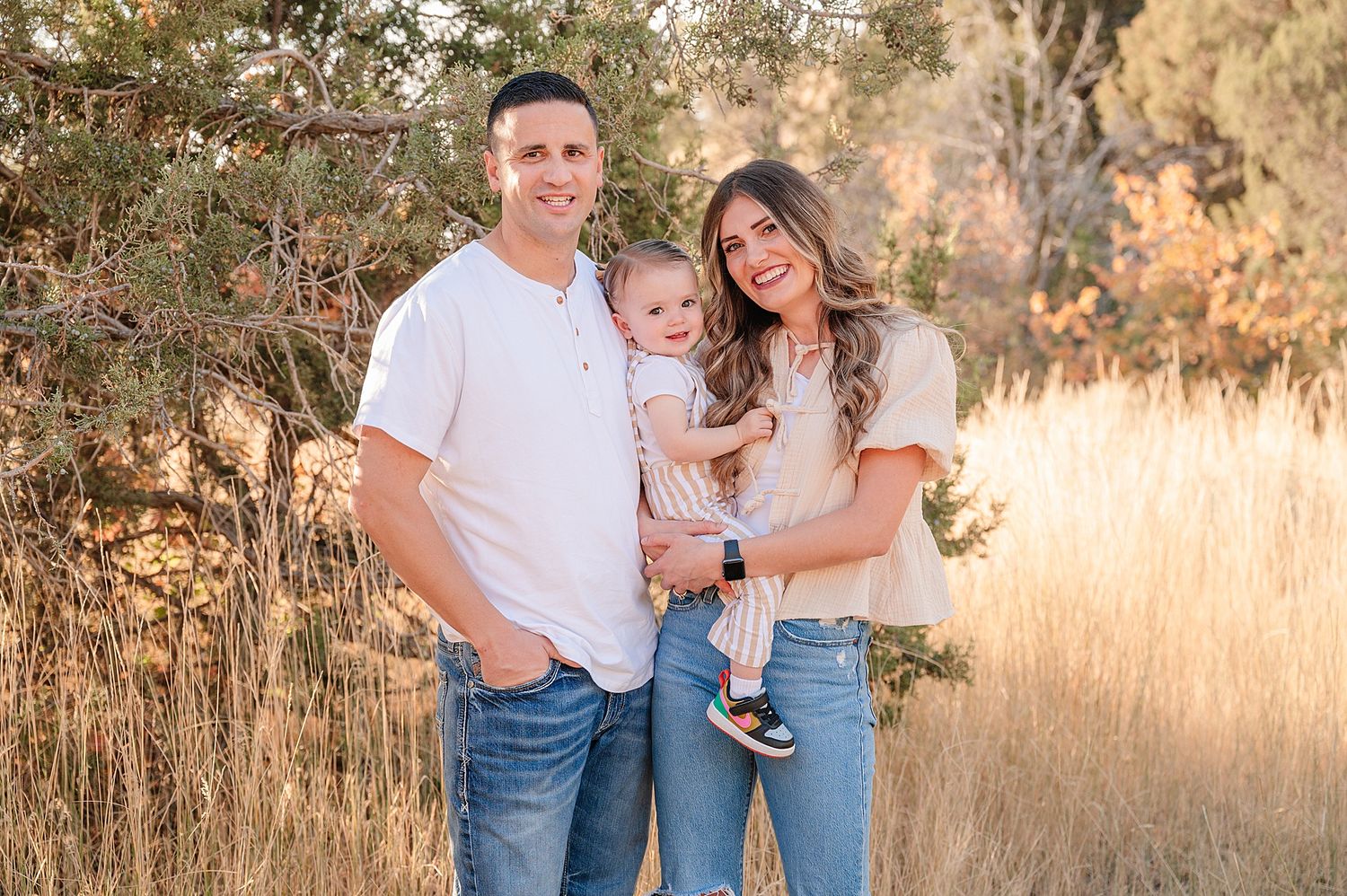Family of three stands together in a field of tall grass at golden hour.