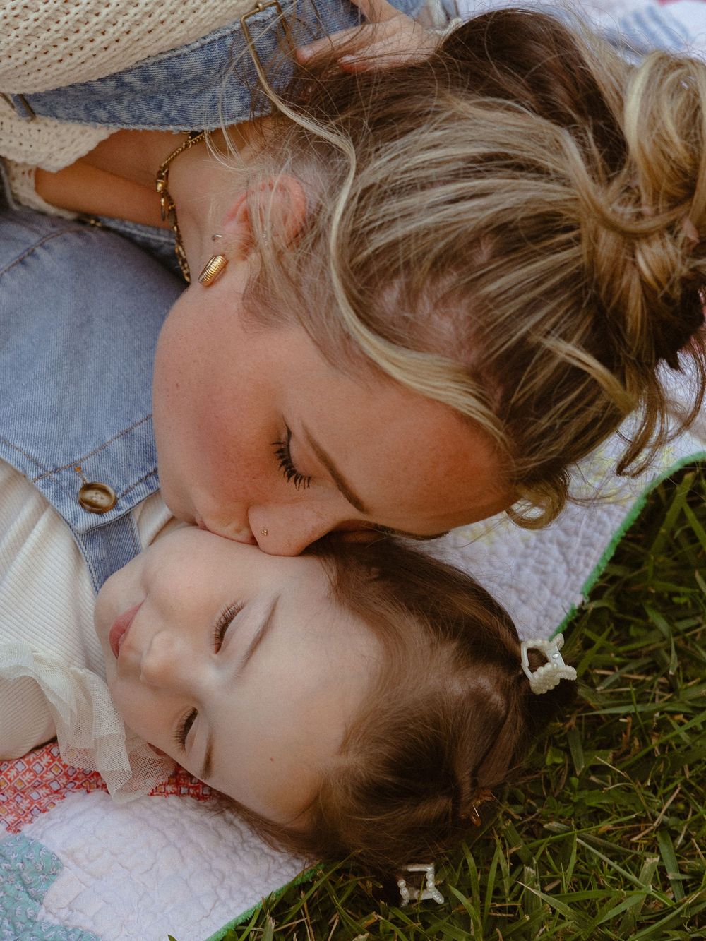 A mother tenderly kisses her baby on the head while lying on a quilt in the grass outdoors.