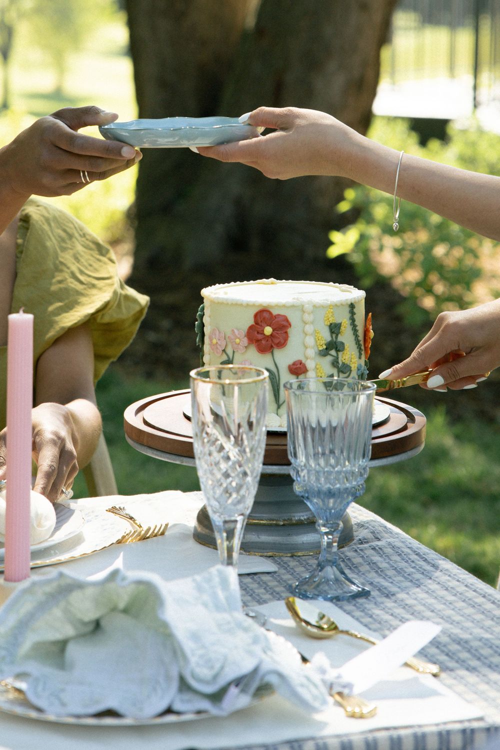 Two people serving slices of a floral decorated cake at an elegant outdoor garden party table setting.