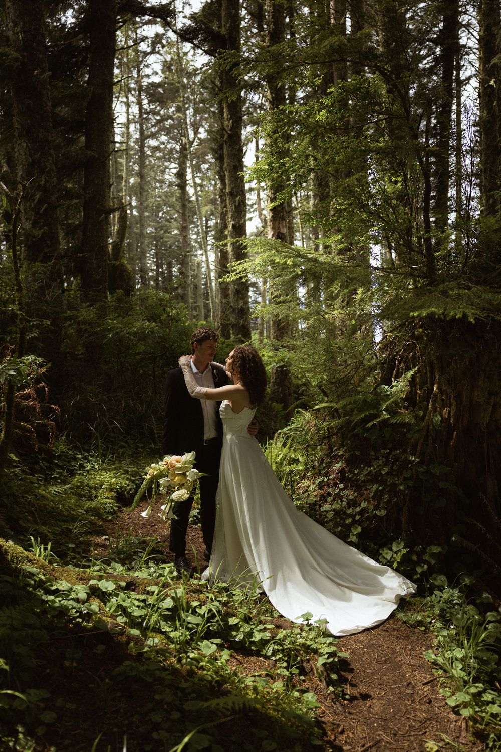 Bride and groom share a romantic moment on a forest path surrounded by lush green ferns and tall trees.