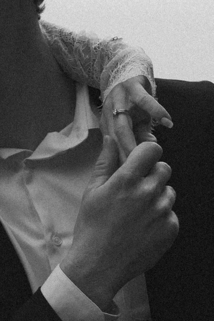 Black and white photo of two hands holding, one wearing a ring and lace sleeve, the other in a suit jacket.