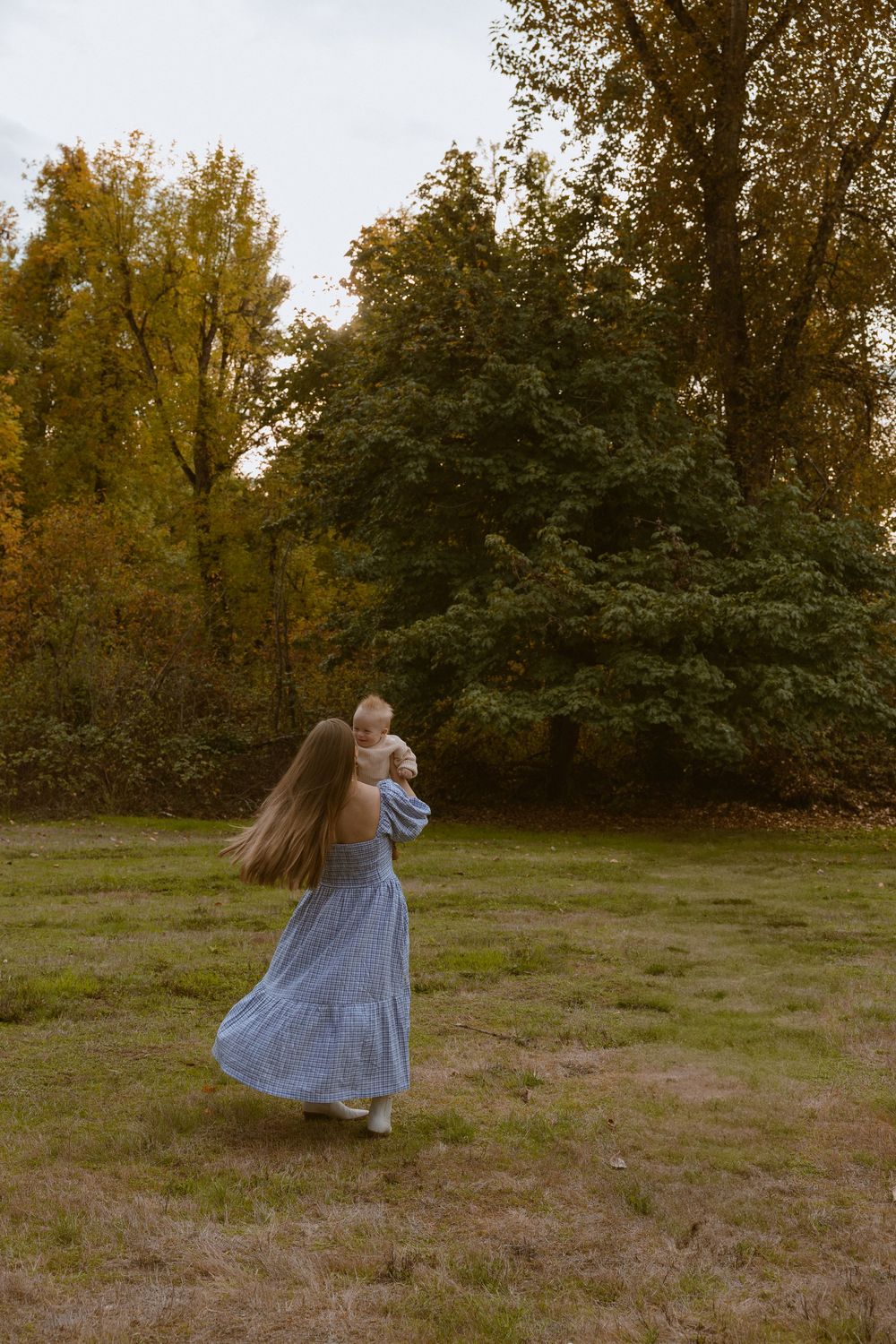 A woman in a blue dress with long hair holds a baby while walking in a lush autumn park.