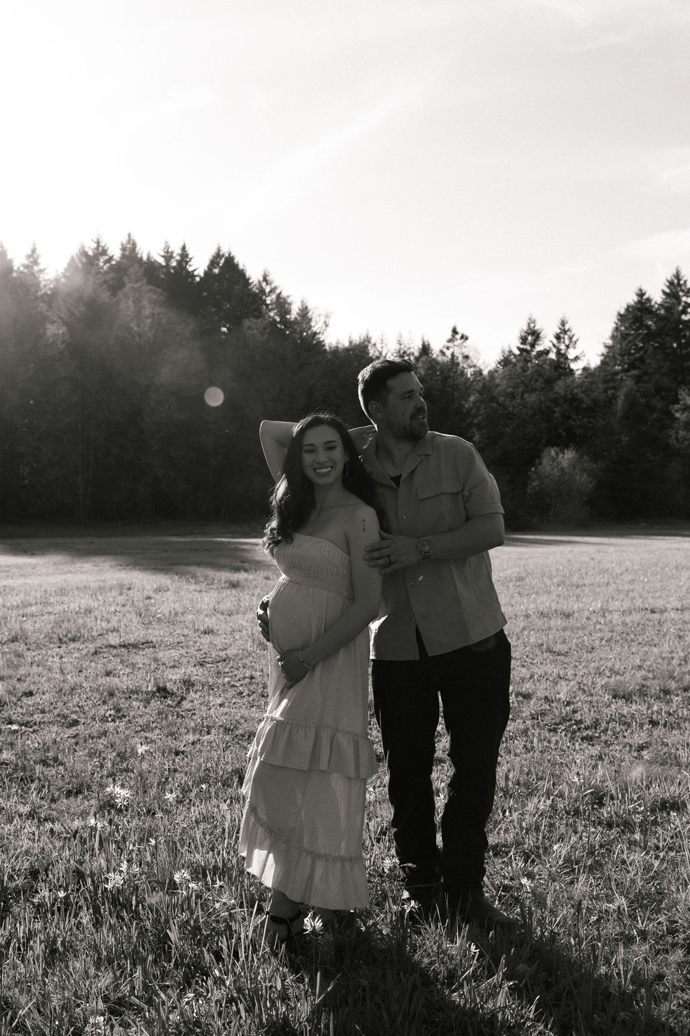 Black and white maternity photo of a couple embracing in a sunlit field with trees in the background.