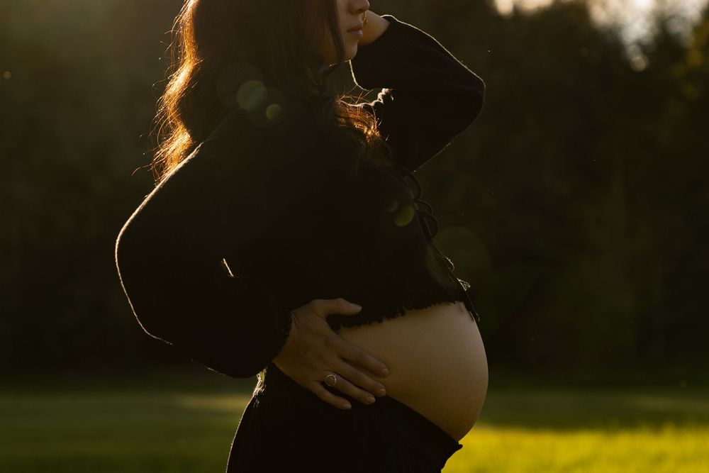 Pregnant woman in black outfit cradling her baby bump outdoors in golden sunset light in a park.