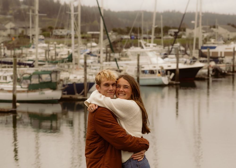 Couple embracing and smiling at a marina with sailboats in the background on a cloudy day.