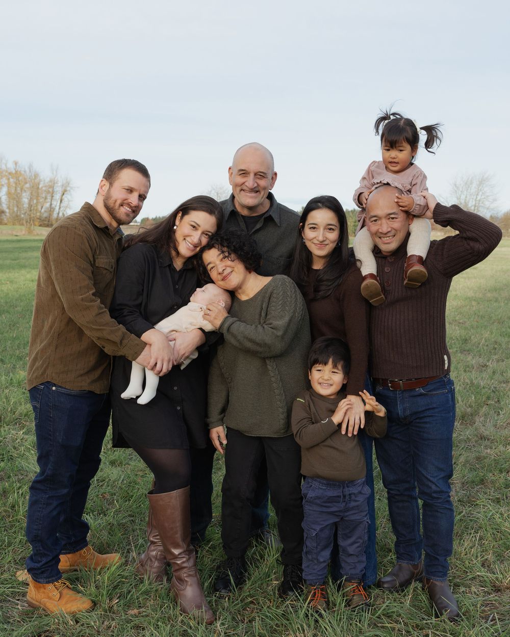 Multi-generational family portrait outdoors on green grass field, smiling together on a cloudy day.