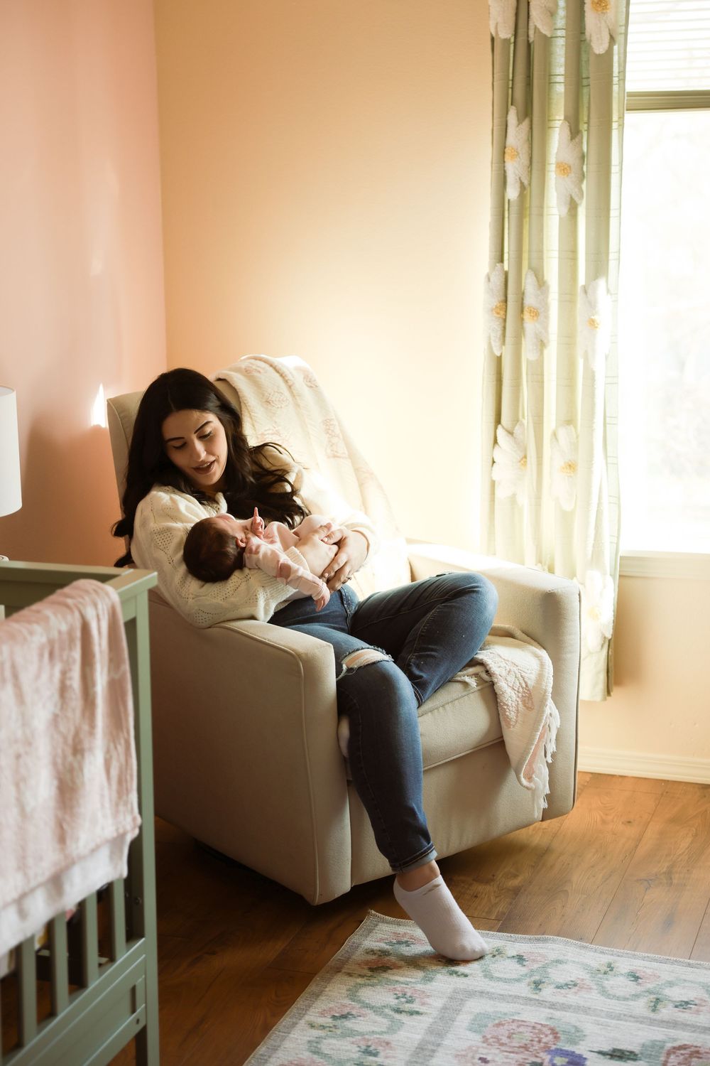Mother sitting in nursery glider chair holding newborn baby, soft pink walls and floral curtains in background.