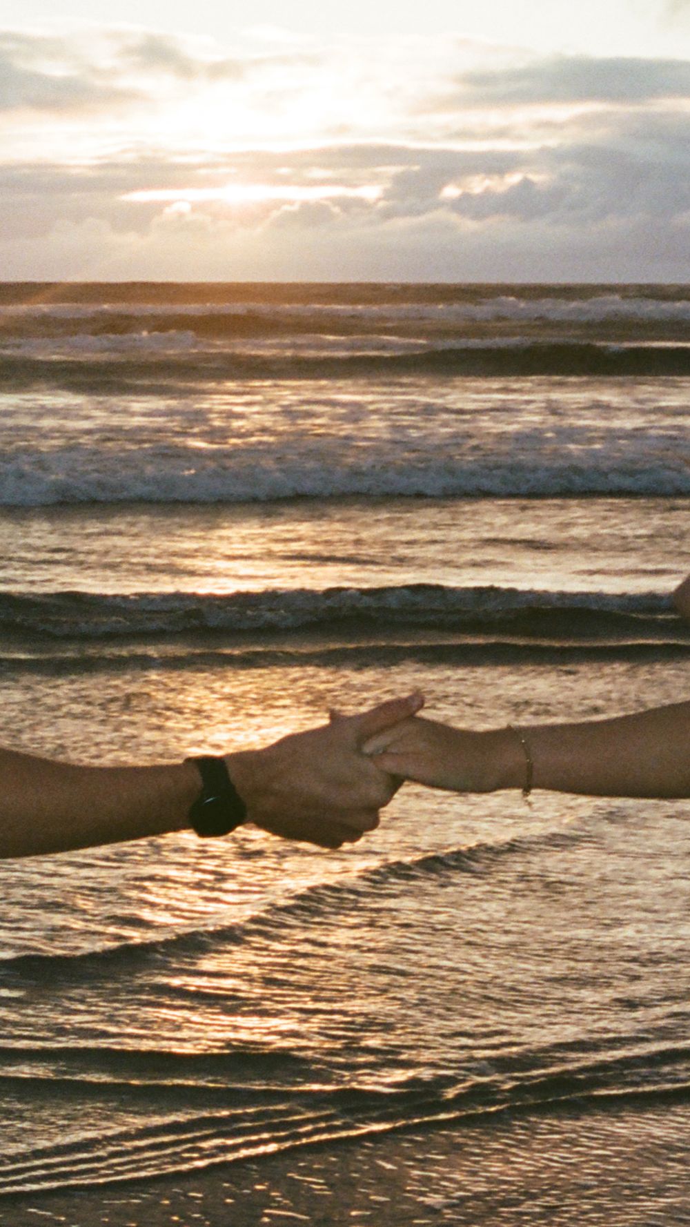 Two hands gently holding fingers at a golden sunset beach with ocean waves in the background.