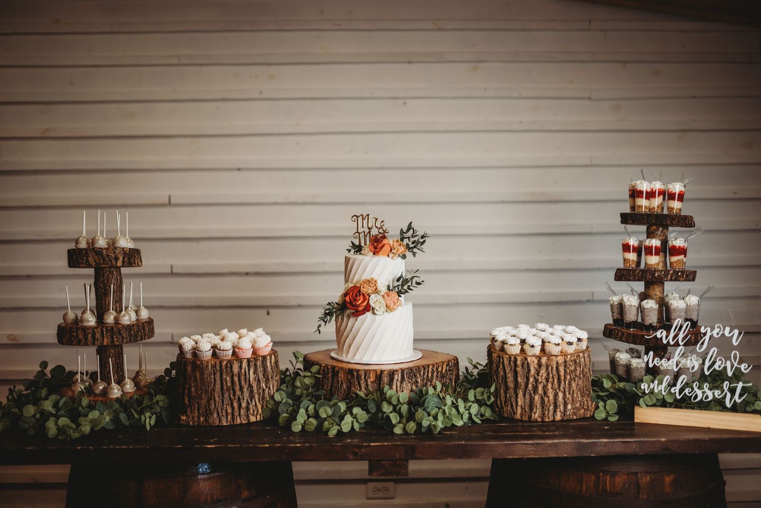 Rustic wedding dessert table with three-tiered cake and wooden log stands decorated with greenery.