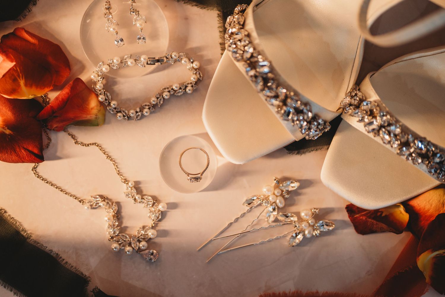 Wedding accessories including jewelry, rings and orange flowers arranged on light fabric.