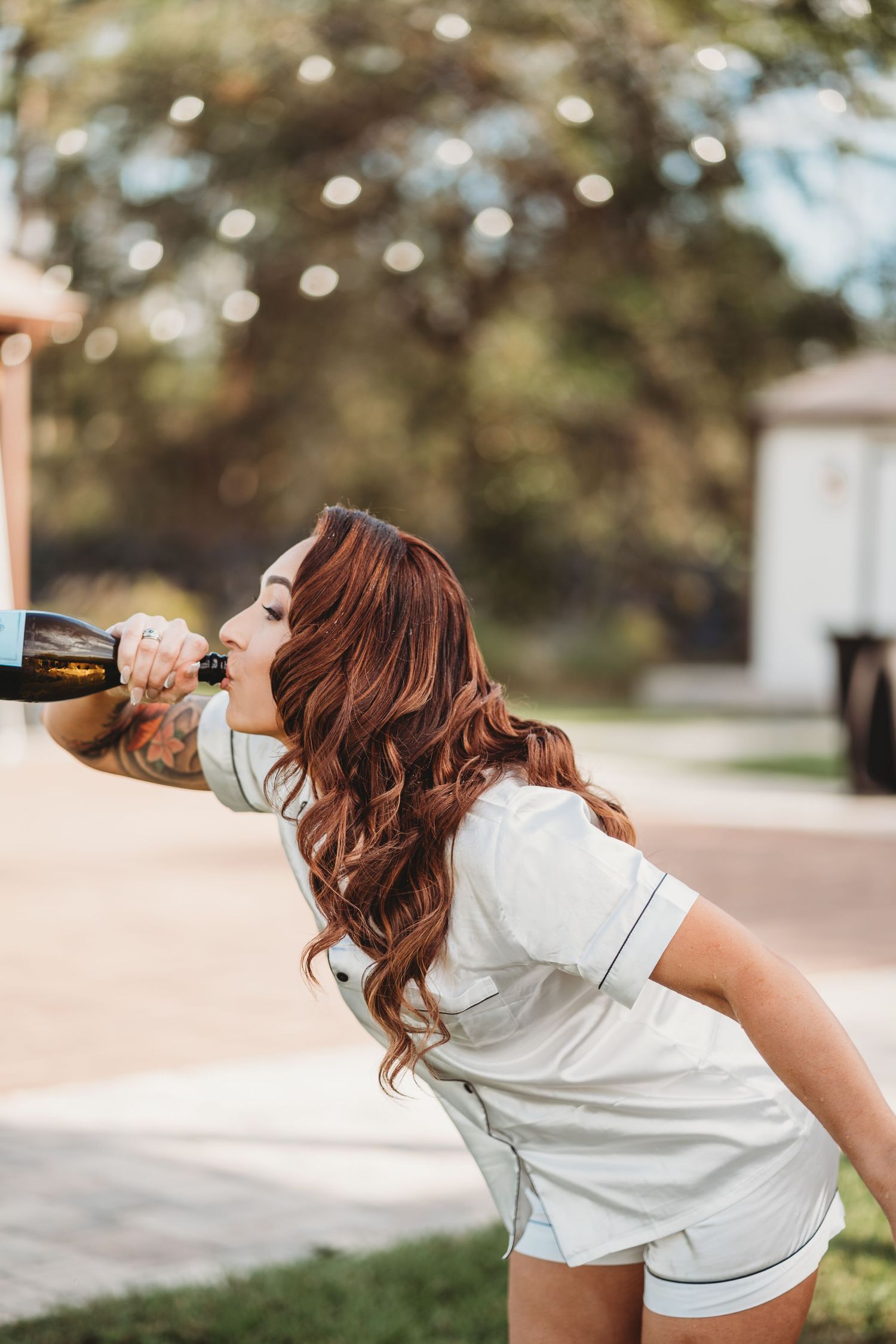 Woman in white outfit drinking from a bottle outdoors with bokeh lights in background.