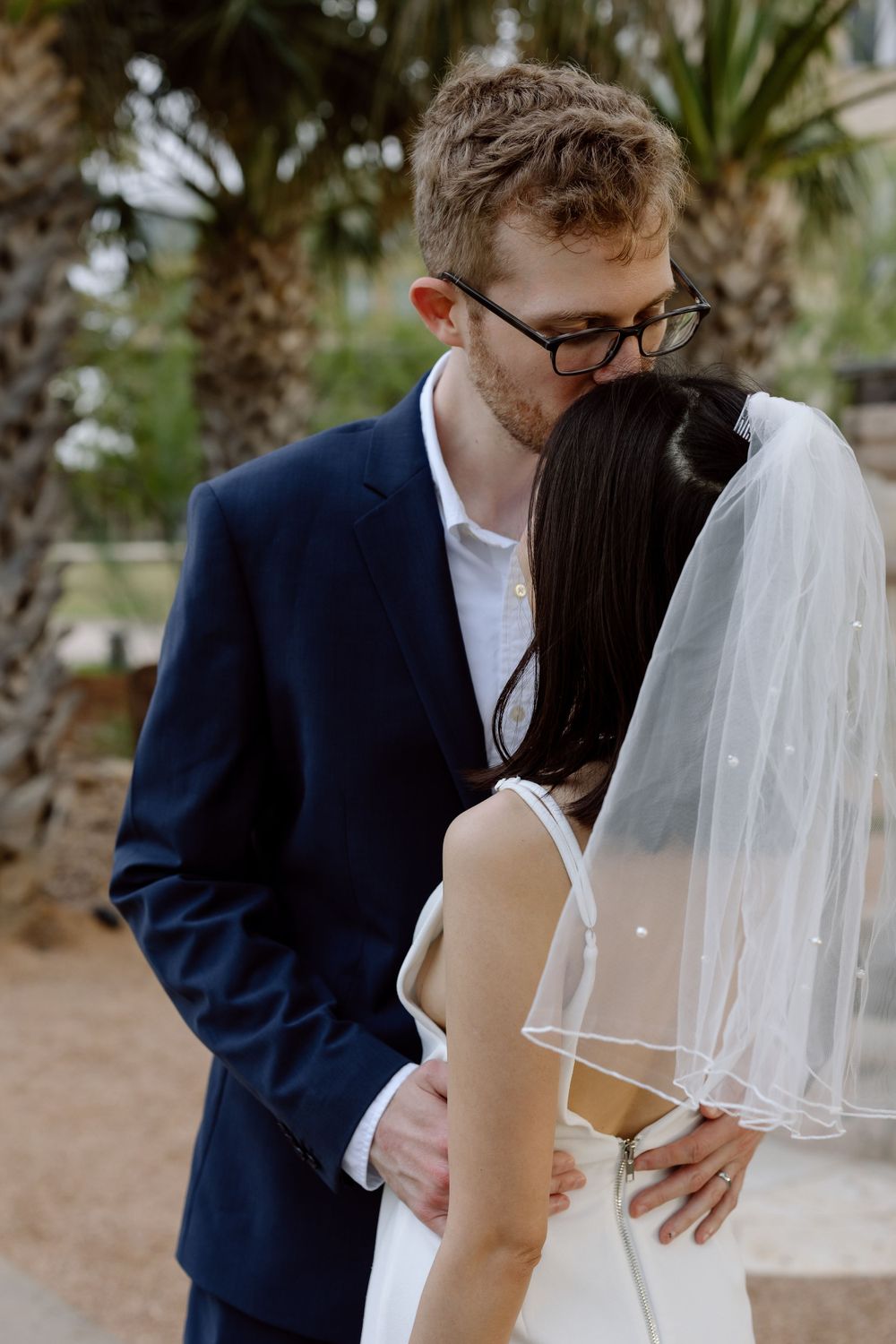 Intimate wedding moment between a couple embracing outdoors with palm trees in the background.