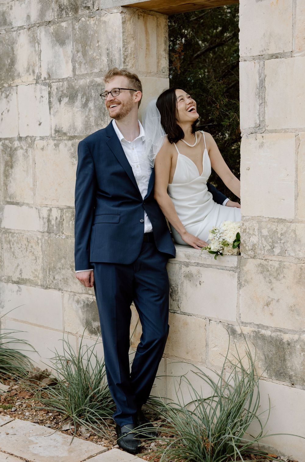 A happy couple in wedding attire laughs while leaning against an outdoor stone wall with ornamental grasses.