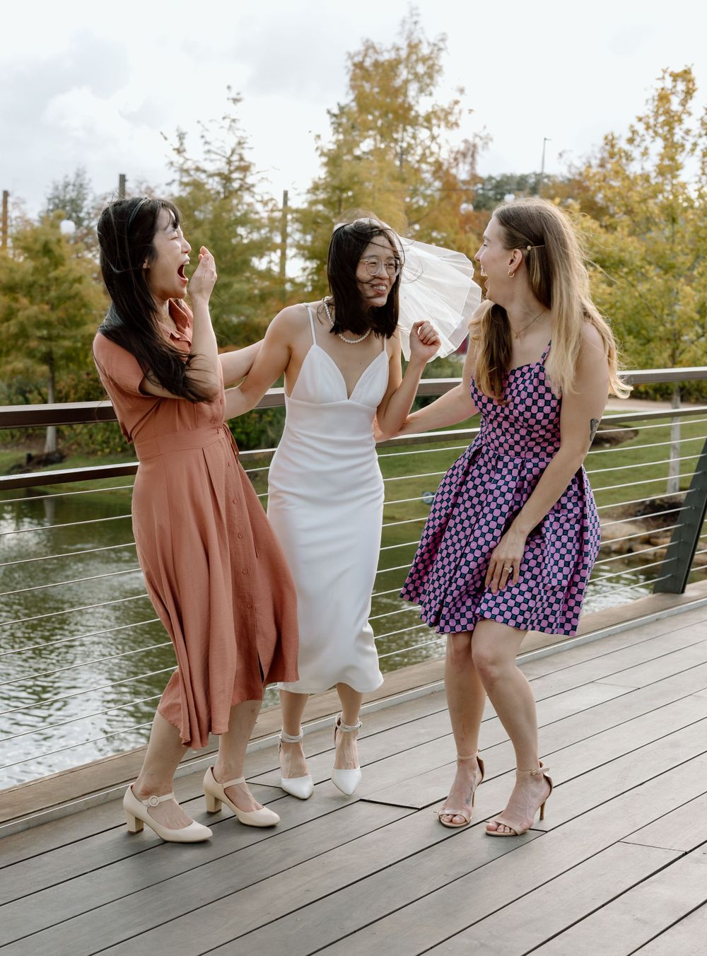 Three friends in flowing dresses laugh and dance together on a wooden deck by a lake during autumn.