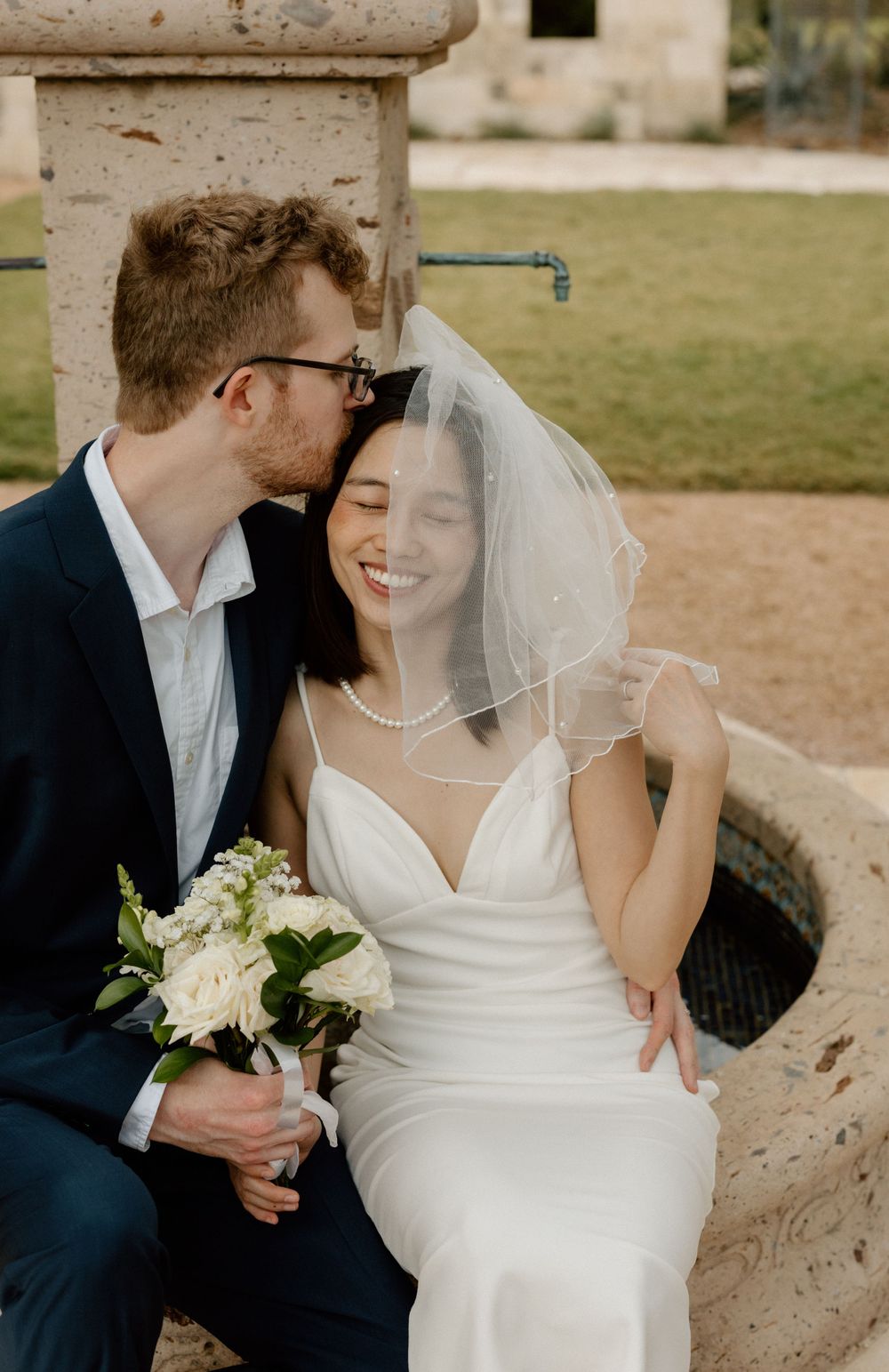 groom kissing bride on forehead while she smiles towards the camera