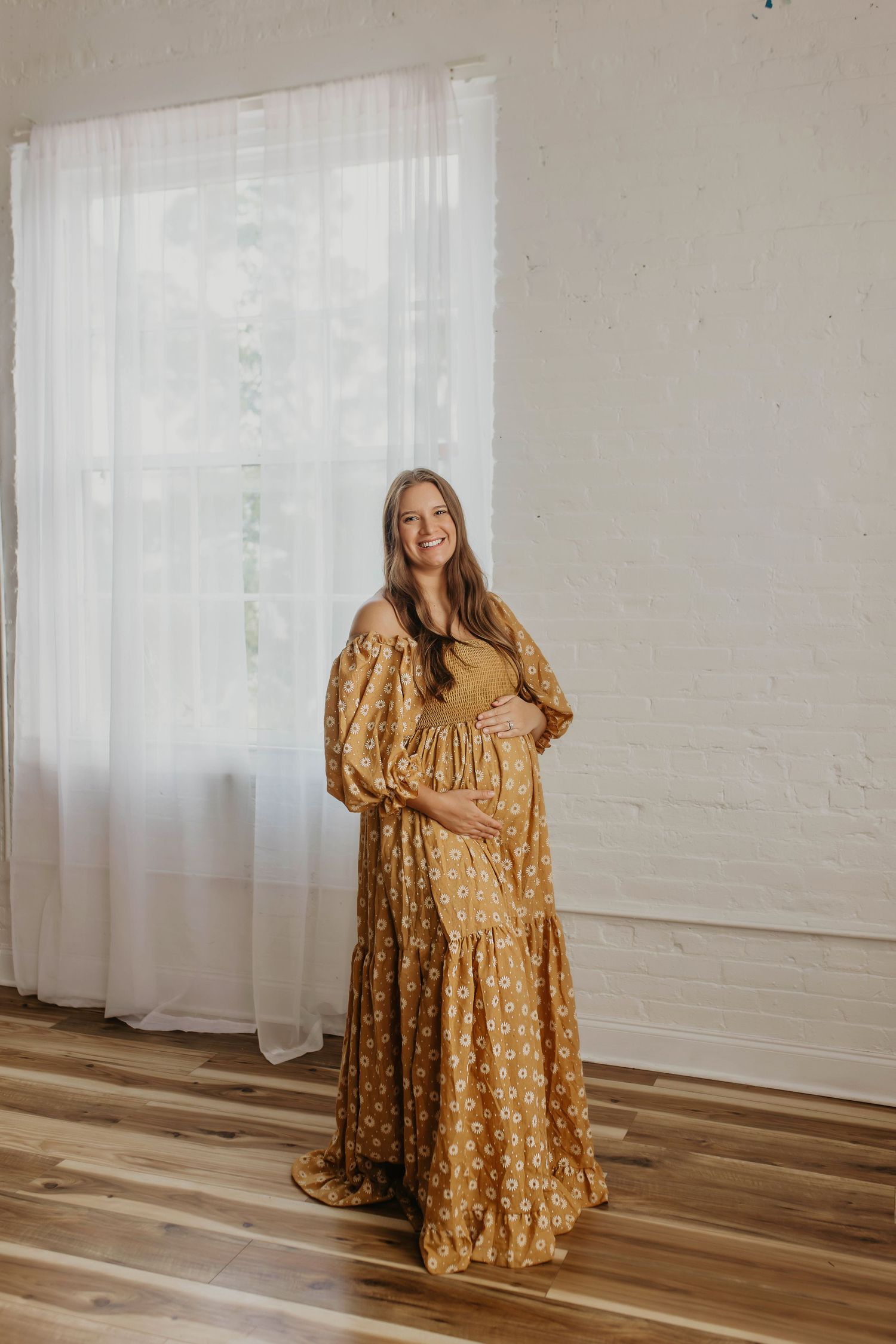 A maternity photo of someone wearing a flowing mustard yellow prairie-style dress in a bright minimalist room.