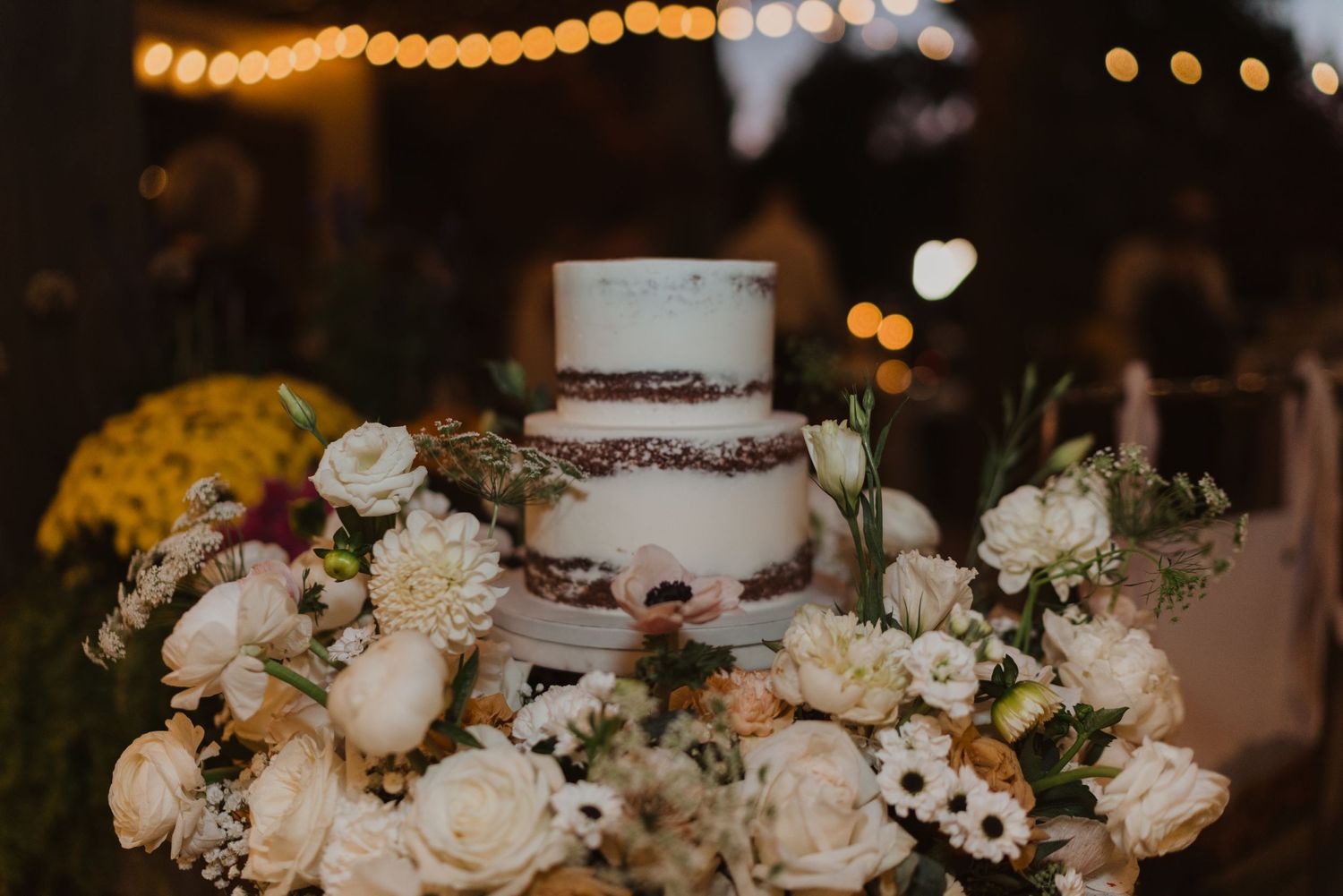 Elegant wedding cake decorated with white roses and string lights in the background.
