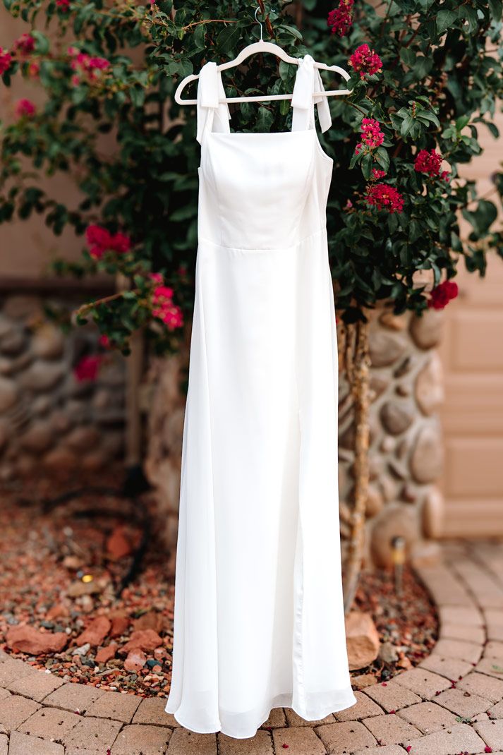 Classic white wedding dress hanging on display against flowering bush with red blooms.