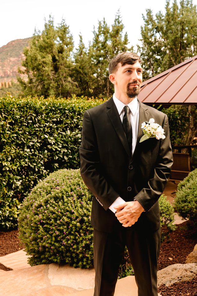 A groom in a classic black suit with boutonniere stands in front of greenery and Sedona mountains on his wedding day.