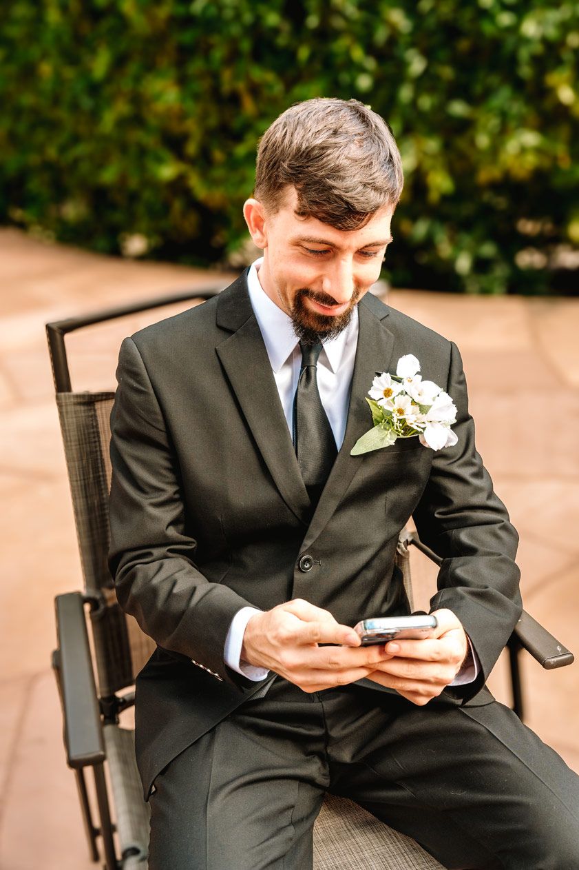 A groom in a black suit checks his phone while seated outdoors on his wedding day.