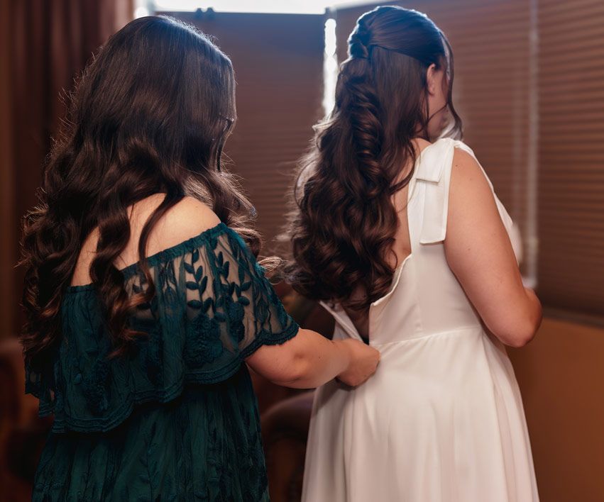 Two people in formal dresses, one teal and one white, shown from behind with elegant curled hairstyles.