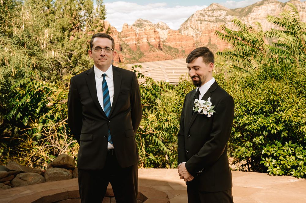 Two men in formal suits stand together during a wedding ceremony with Sedona red rock mountains in the background.