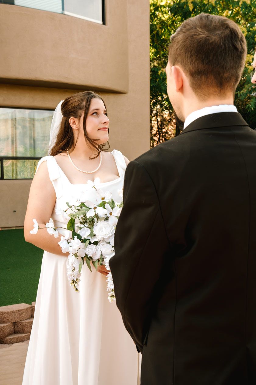Bride in white dress holds bouquet while facing groom during outdoor ceremony.