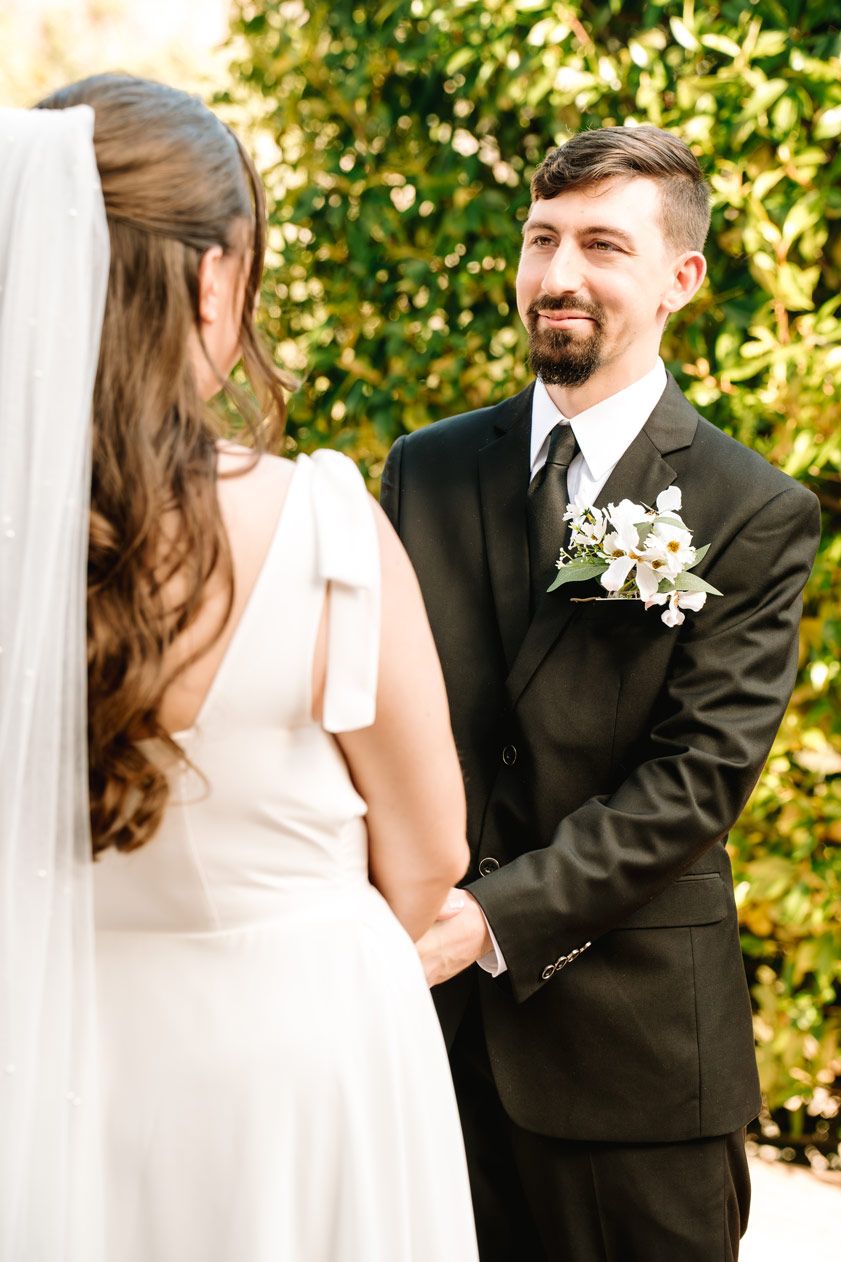 A wedding ceremony moment between a bride in white and groom in black suit against green foliage.