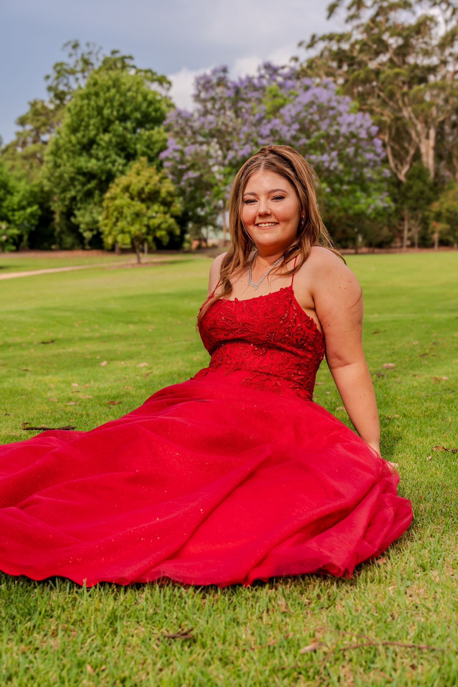 A woman in a flowing red formal dress sits gracefully on lush green grass with blooming trees in the background.