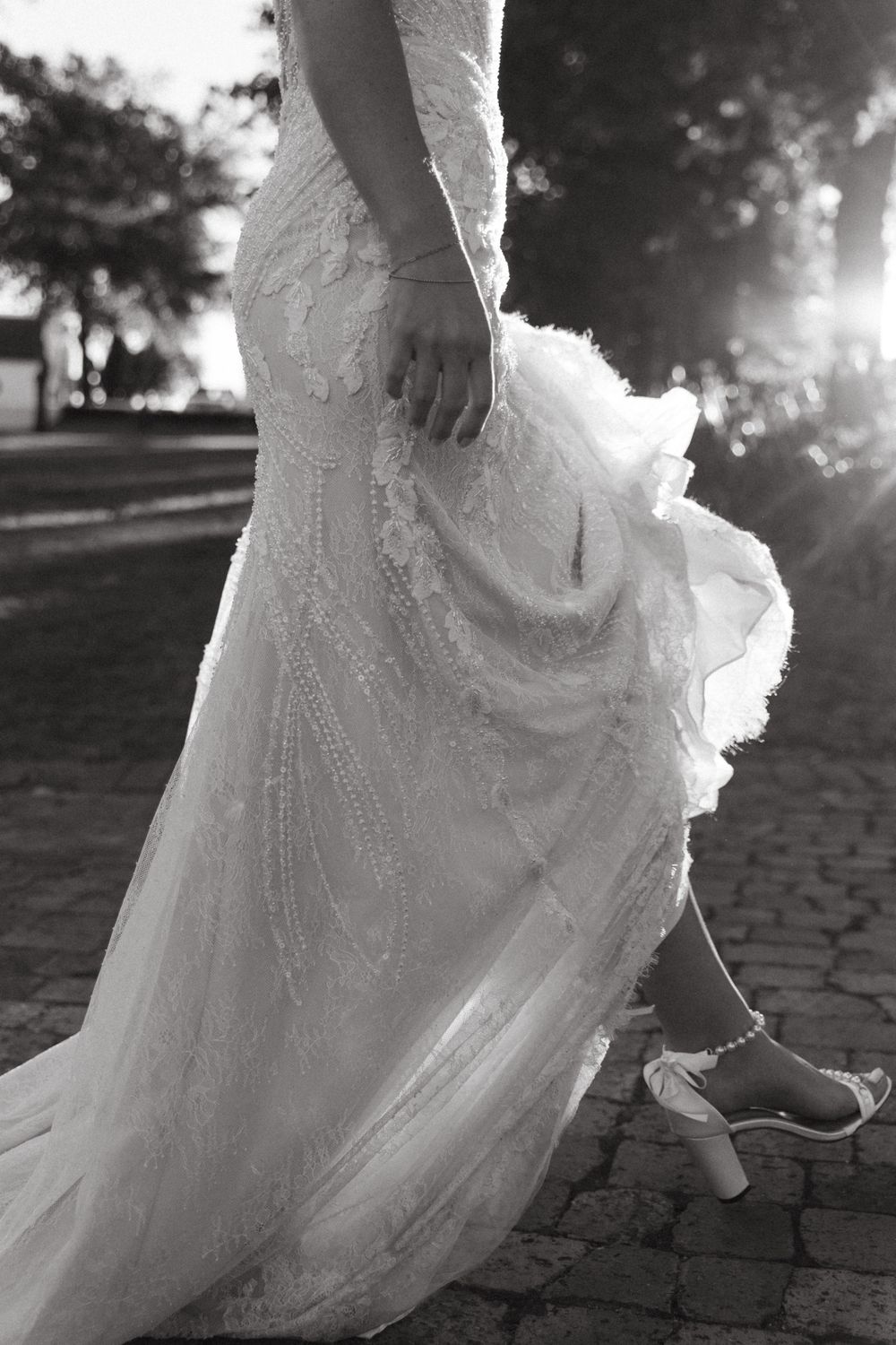 Detail shot of an elegant lace wedding dress train catching sunlight on cobblestone path.