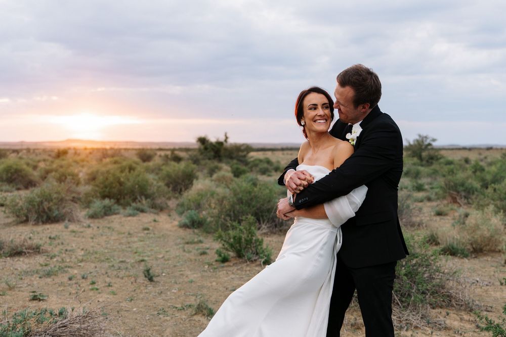 A couple in wedding attire embraces in a desert landscape at sunset with mountains in the background.