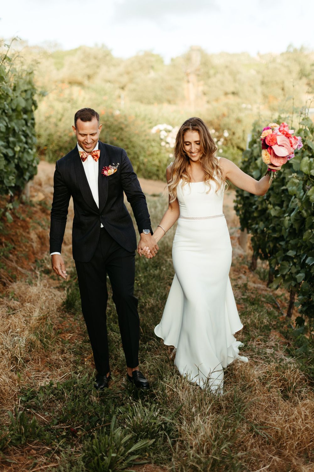 A couple walking hand in hand through a vineyard, the bride in white and groom in black.
