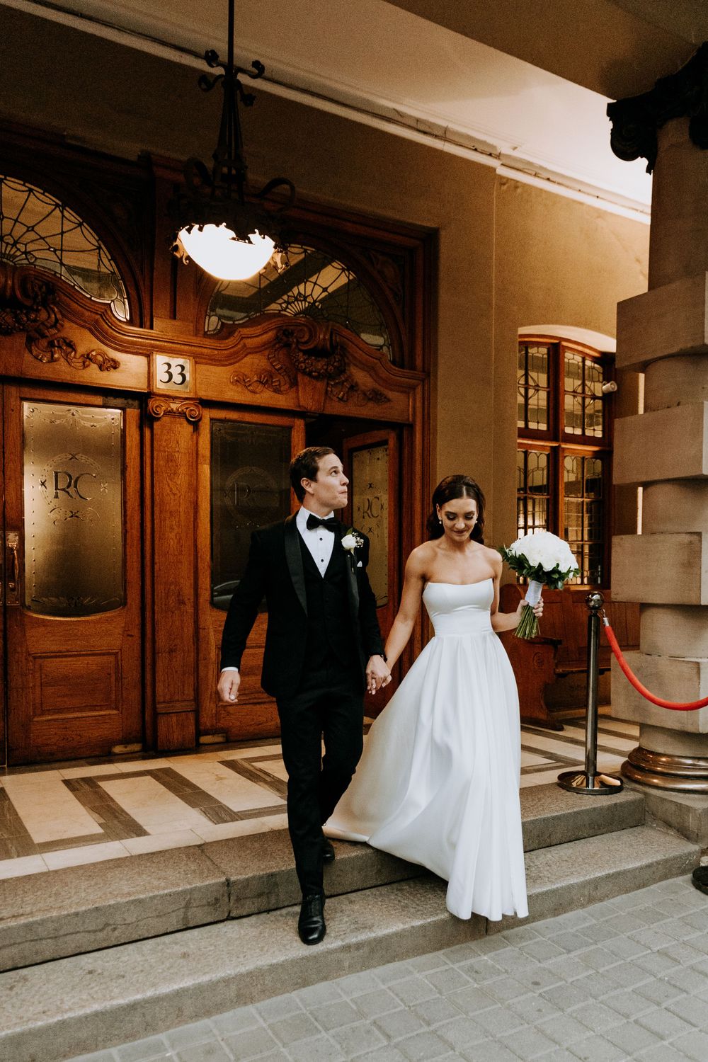 A wedding couple exits an ornate vintage building with wooden doors and architectural details.
