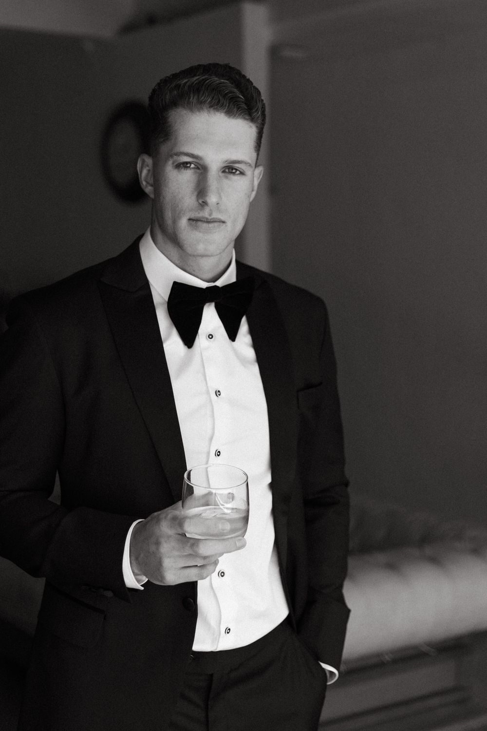 Black and white photo of an elegant man in a tuxedo with bow tie holding a glass drink.