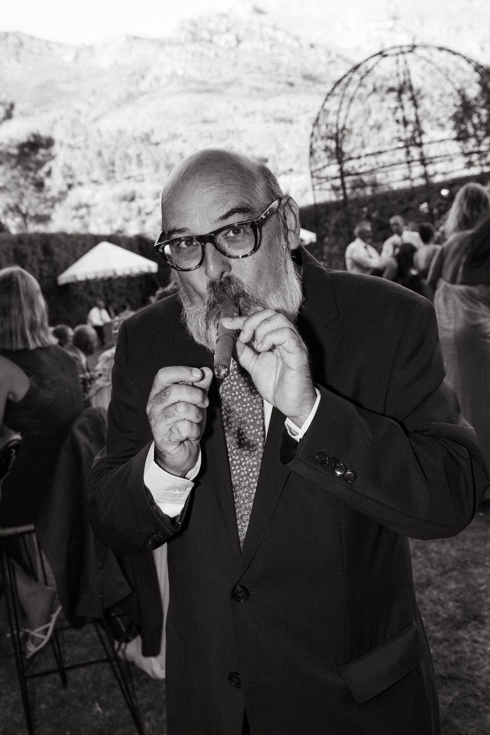 Bald man with goatee in suit and glasses twirling mustache at outdoor garden party, black and white photo.