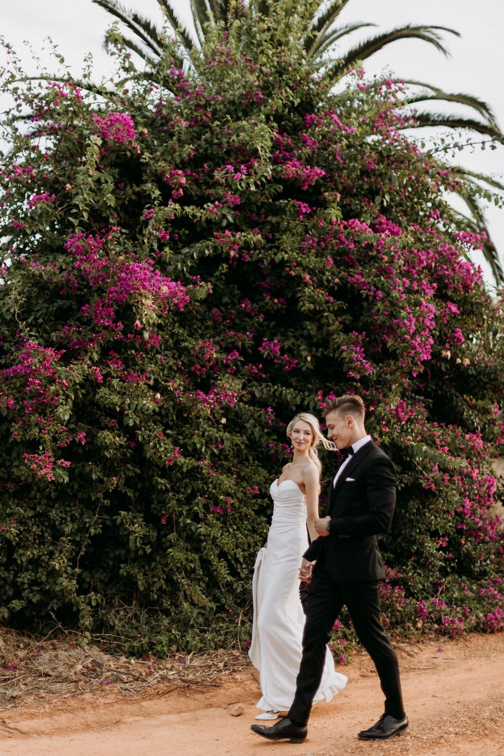A romantic wedding couple walking on a path beneath vibrant pink bougainvillea and palm trees.