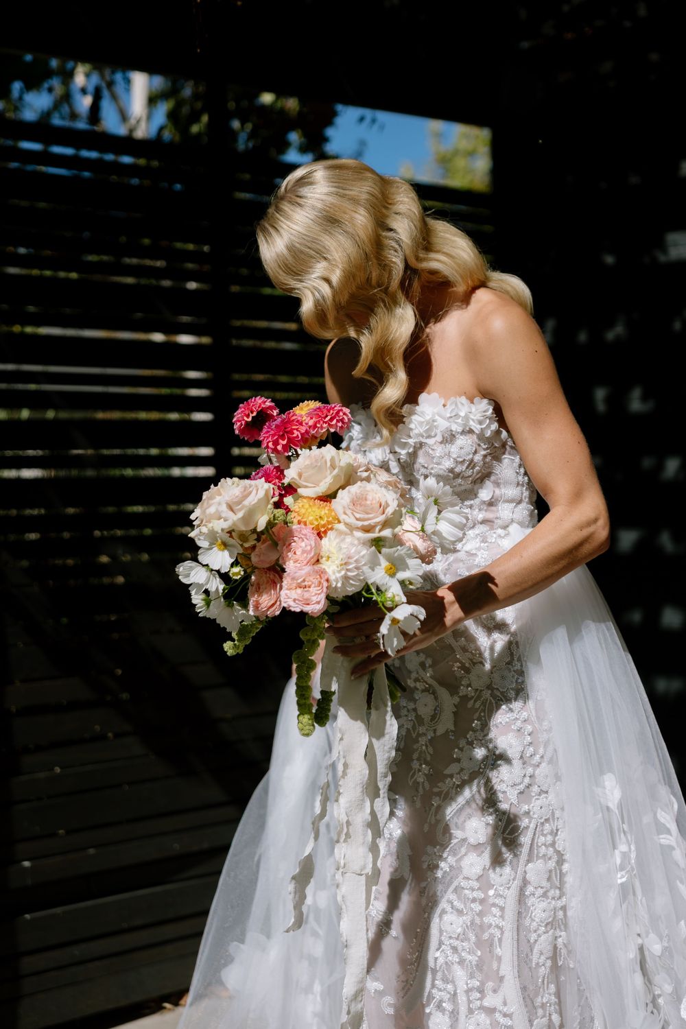 Bride in strapless lace wedding gown holding colorful bouquet of pink and white flowers, head bowed in sunlight.